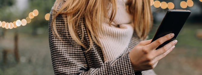 A woman in a coat and scarf walking in a city while holding a cup of coffee and reading her mobile phone.